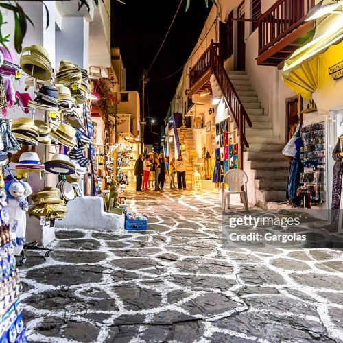 Mykonos, Greece - September 21, 2013:  In the alley of stone or everything is white, there are merchants of various souvenirs. They are open during the day and especially at night to accommodate customers looking for nightlife as good food and dancing. The displays are arranged along the lane and attract customers by the colors and diversity. We see tourists in the streets discussing and walking.